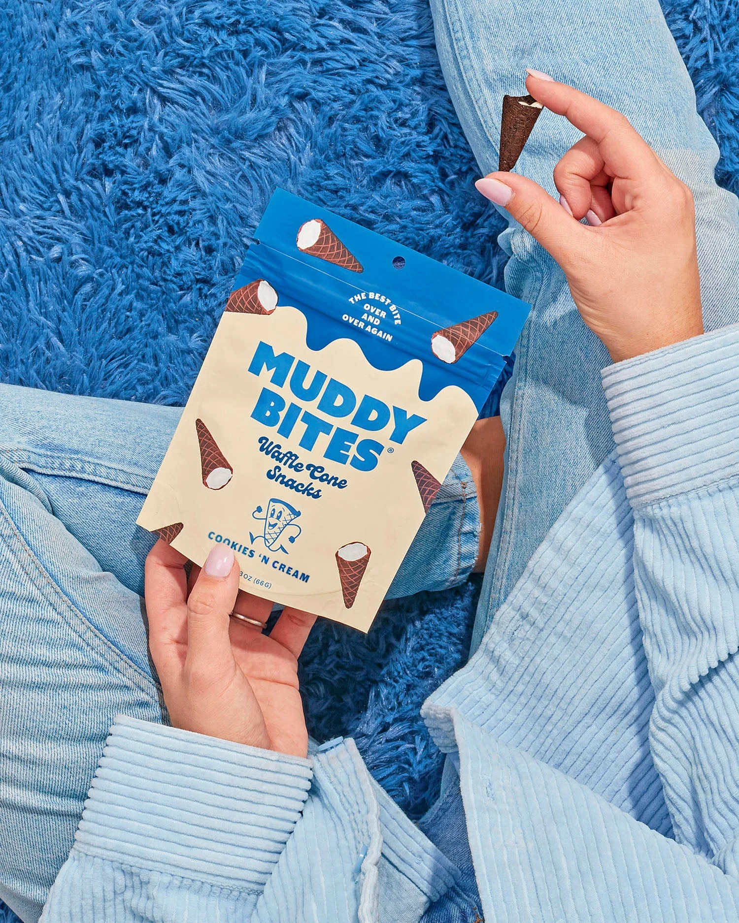 A hand holds a bag of Muddy Bites Cookies 'N Cream over a blue rug